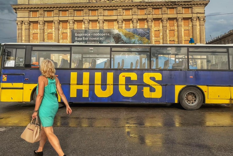 A woman passes a bus in central Kharkiv on June 12. Behind, the Kharkiv city hall displays a banner reading, "Fight, you will win — God will help you!"