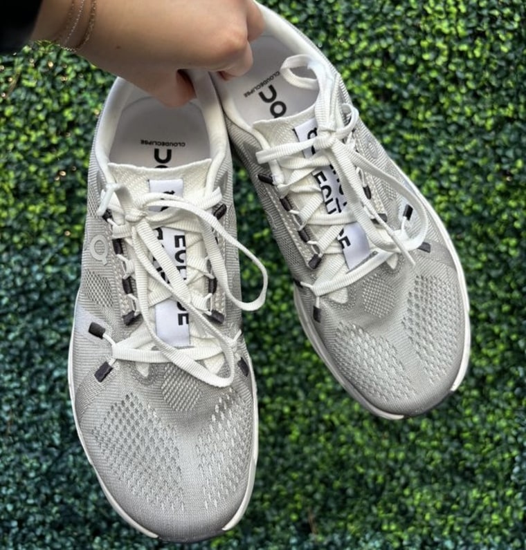 Woman's hand holding a pair of grey and white On Cloudeclipse sneakers against a green background.