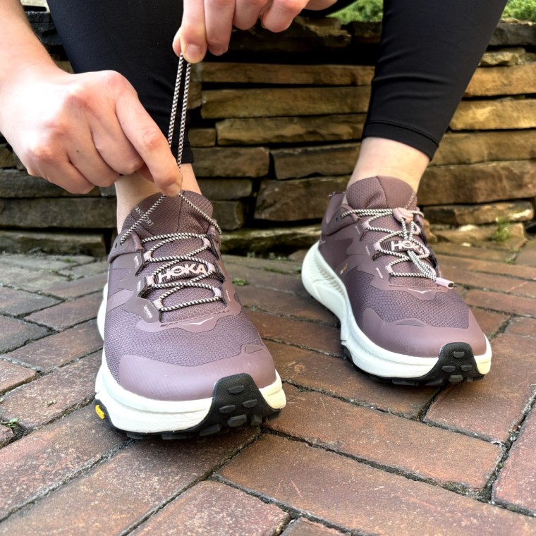 A woman sitting against a stone wall, pulling the elastic laces tighter on the Hoka Transport sneaker.